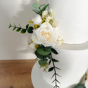 K&ouml;nnte beinhalten: Eine zweist&ouml;ckige wei&szlig;e Hochzeitstorte, die mit einer kaskadierenden Anordnung von wei&szlig;en Rosen und Gr&uuml;n verziert ist.
