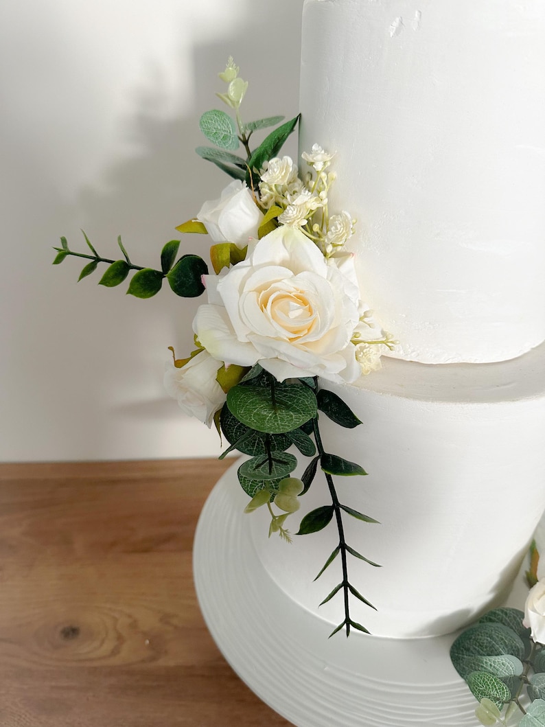 K&ouml;nnte beinhalten: Eine zweist&ouml;ckige wei&szlig;e Hochzeitstorte, die mit einer kaskadierenden Anordnung von wei&szlig;en Rosen und Gr&uuml;n verziert ist.