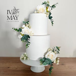 K&ouml;nnte beinhalten: Zweist&ouml;ckige wei&szlig;e Hochzeitstorte, verziert mit wei&szlig;en Rosen und Gr&uuml;n. Die Torte steht auf einem wei&szlig;en Tortenst&auml;nder.