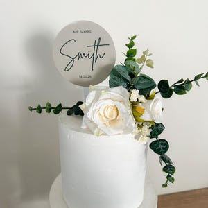 K&ouml;nnte beinhalten: Eine zweist&ouml;ckige wei&szlig;e Hochzeitstorte mit einem runden silbernen Topper, auf dem "Mr. & Mrs. Smith 14.02.26" steht. Die Torte ist mit wei&szlig;en Rosen und Gr&uuml;n verziert.