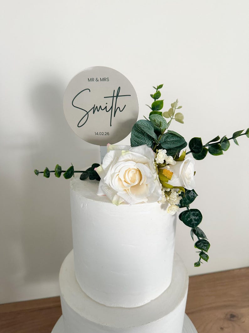 K&ouml;nnte beinhalten: Eine zweist&ouml;ckige wei&szlig;e Hochzeitstorte mit einem runden silbernen Topper, auf dem "Mr. & Mrs. Smith 14.02.26" steht. Die Torte ist mit wei&szlig;en Rosen und Gr&uuml;n verziert.