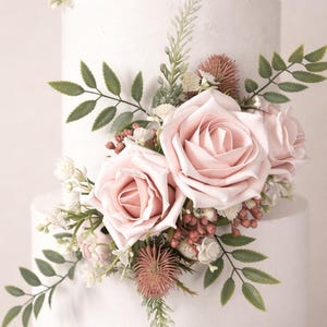 Op de afbeelding: Een witte taart met twee lagen, versierd met roze rozen, groene bladeren en kleine witte en rode bloemen. De taart heeft een getextureerd oppervlak, ideaal voor een bruiloft of een speciale gelegenheid.