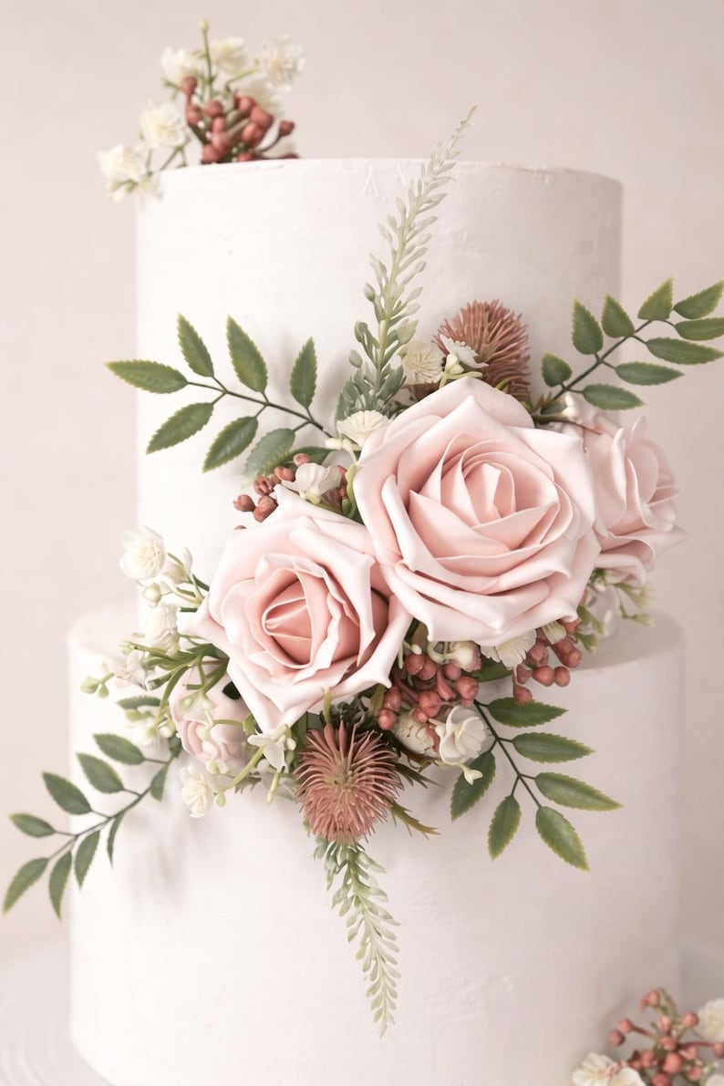 Op de afbeelding: Een witte taart met twee lagen, versierd met roze rozen, groene bladeren en kleine witte en rode bloemen. De taart heeft een getextureerd oppervlak, ideaal voor een bruiloft of een speciale gelegenheid.