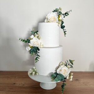 K&ouml;nnte beinhalten: Zweist&ouml;ckige wei&szlig;e Hochzeitstorte, verziert mit wei&szlig;en Rosen und Gr&uuml;n.