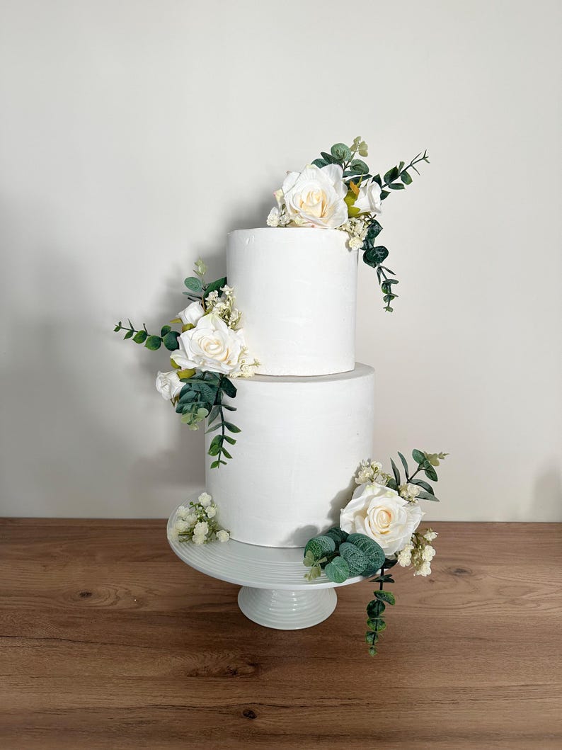 K&ouml;nnte beinhalten: Zweist&ouml;ckige wei&szlig;e Hochzeitstorte, verziert mit wei&szlig;en Rosen und Gr&uuml;n.