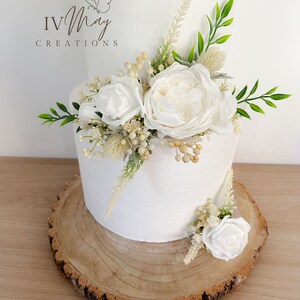 Op de afbeelding: Een witte taart met twee lagen, versierd met witte bloemen, groen en gedroogde bloemaccenten. De taart staat op een rustieke houten schijf. Het logo "IV May Creations" is zichtbaar.