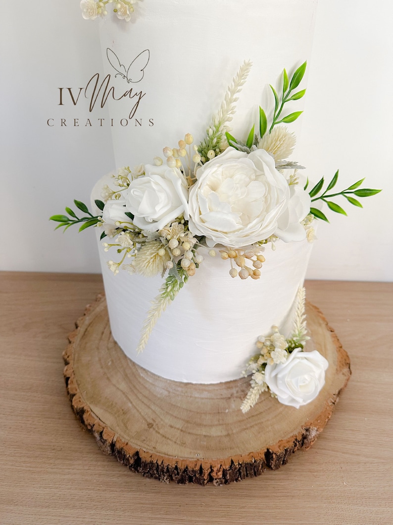 Op de afbeelding: Een witte taart met twee lagen, versierd met witte bloemen, groen en gedroogde bloemaccenten. De taart staat op een rustieke houten schijf. Het logo "IV May Creations" is zichtbaar.