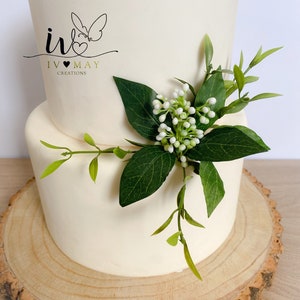 May include: A two-tiered cake with smooth off-white frosting, decorated with a cluster of green leaves and small white berries. The cake features a logo with a butterfly and the text IV MAY CREATIONS. It is displayed on a round wooden slice.