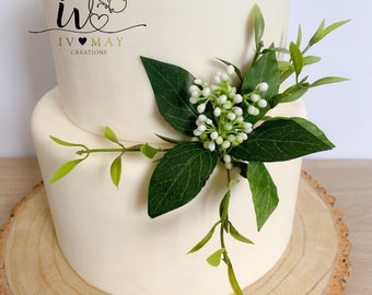 May include: A two-tiered cake with smooth off-white frosting, decorated with a cluster of green leaves and small white berries. The cake features a logo with a butterfly and the text IV MAY CREATIONS. It is displayed on a round wooden slice.