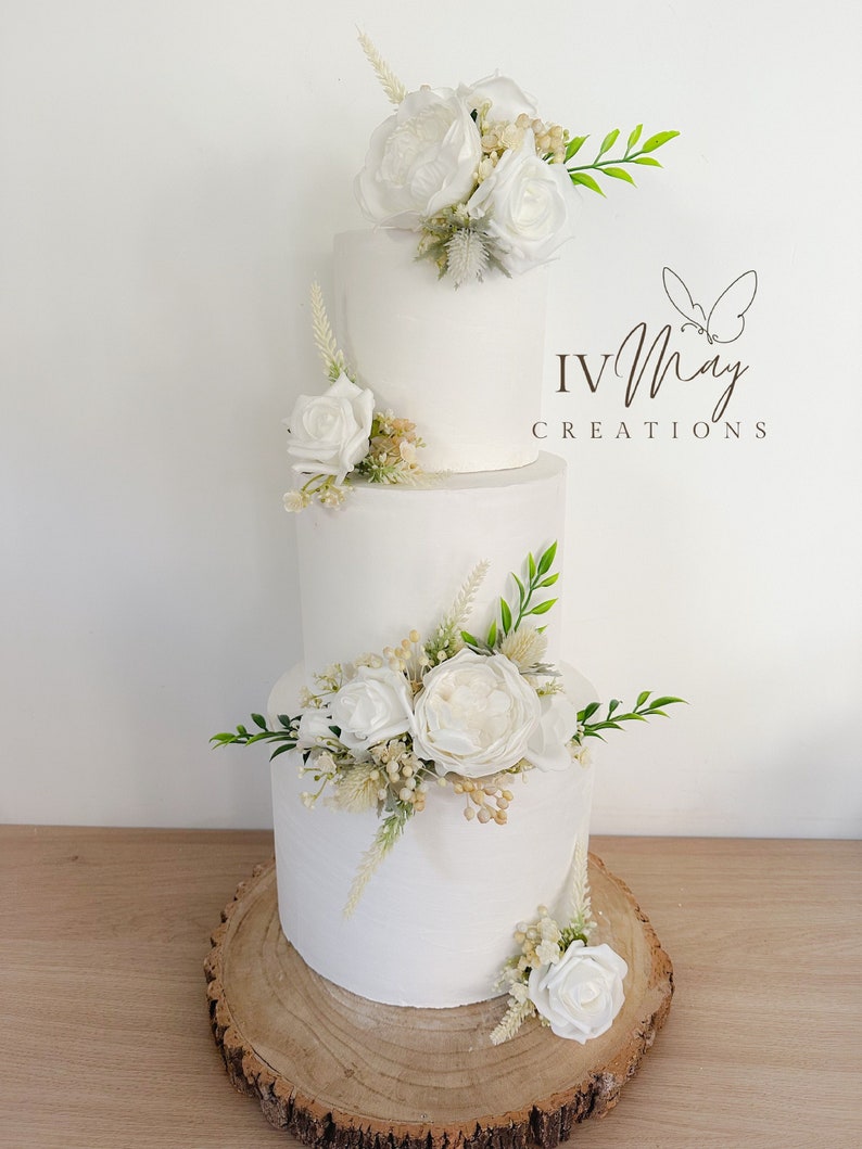 Op de afbeelding: Een witte taart met drie lagen, versierd met witte rozen, groen en bloemaccenten. De taart staat op een houten schijf. De tekst "IV May Creations" is zichtbaar aan de rechterkant van de taart.