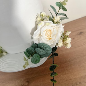 K&ouml;nnte beinhalten: Ein wei&szlig;er Tortendekor mit einer einzelnen wei&szlig;en Rose, kleinen wei&szlig;en Bl&uuml;ten und gr&uuml;nen Eukalyptusbl&auml;ttern.