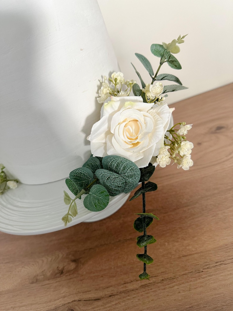 K&ouml;nnte beinhalten: Ein wei&szlig;er Tortendekor mit einer einzelnen wei&szlig;en Rose, kleinen wei&szlig;en Bl&uuml;ten und gr&uuml;nen Eukalyptusbl&auml;ttern.