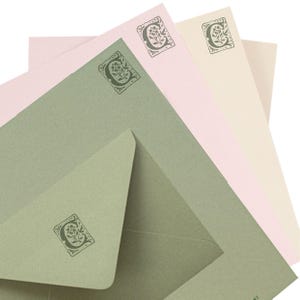 May include: A collection of stationery featuring sage green, light pink, and cream-coloured paper and envelopes. Each piece is decorated with a dark green rectangular stamp displaying the letter C surrounded by a floral and leaf botanical design.