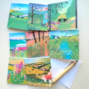 May include: A collection of landscape art greeting cards depicting rural scenes with fields, flowers, and a pink campervan. A wooden pen and white envelopes are also included.