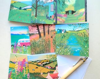 May include: A set of landscape-themed greeting cards featuring colorful illustrations of rural scenes, including fields, flowers, and a pink van. A wooden pen and white envelopes are also included.