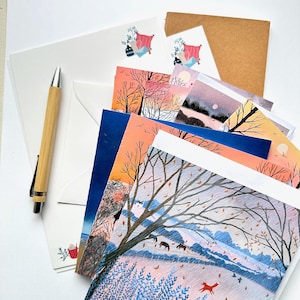 May include: A collection of stationery featuring various landscape illustrations, including a scene with trees, horses, and a fox, alongside plain white envelopes, a brown envelope, and a bamboo-cased pen with a silver clip and tip.