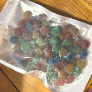 Puede incluir: Una bolsa de plástico transparente sellada llena de caramelos de goma coloridos y azucarados. Los caramelos son de varios colores, incluyendo rojo, verde, azul y amarillo. La bolsa está sobre una superficie de madera.