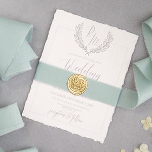 May include: A wedding invitation with a mint green ribbon and gold wax seal. The invitation has a floral wreath design and the word "Wedding" in script. Several pieces of mint green ribbon and small white flowers are arranged around the invitation.