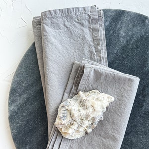 May include: Two gray linen napkins folded on a round gray marble surface. A white seashell is placed on top of the napkins.