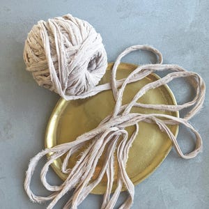 Op de afbeelding: Een bol beige stoffen garen ligt op een goudkleurige schaal, met strengen van dezelfde stof die eruit komen. De schaal staat op een grijsblauwe ondergrond. Het garen lijkt handgemaakt en de algehele esthetiek is rustiek en artistiek.