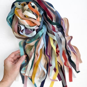 May include: A collection of colorful fabric ribbons in various shades of blue, green, orange, yellow, pink, and purple. The ribbons are arranged in a circular shape, with some strands cascading downwards. The ribbons are held by a hand.