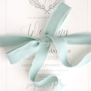 May include: A light cream-colored wedding invitation with a mint green ribbon tied in a bow. The invitation features elegant calligraphy and a decorative wreath with the initials "RM". The text reads "You are invited to Wedding".