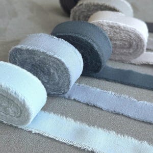 May include: A collection of rolled ribbons in various shades of blue and gray. The ribbons have a frayed edge, giving them a rustic appearance. The colors range from light blue to a deep gray, creating a soft, muted palette.