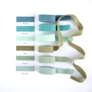 Peut inclure: Rubans en tissu de différentes nuances de bleu, vert et olive, avec des bords effilochés. Un nuancier affiche les noms : Bleu Ardoise, Sauge Bleue, Menthe, Olive, Verdure et Sauge Poussiéreuse. Un ruban olive est également présent.