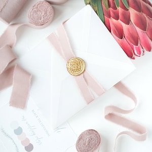 May include: A white envelope with a pink ribbon and a gold wax seal. The envelope is on a white surface with other pink ribbons and a pink flower.