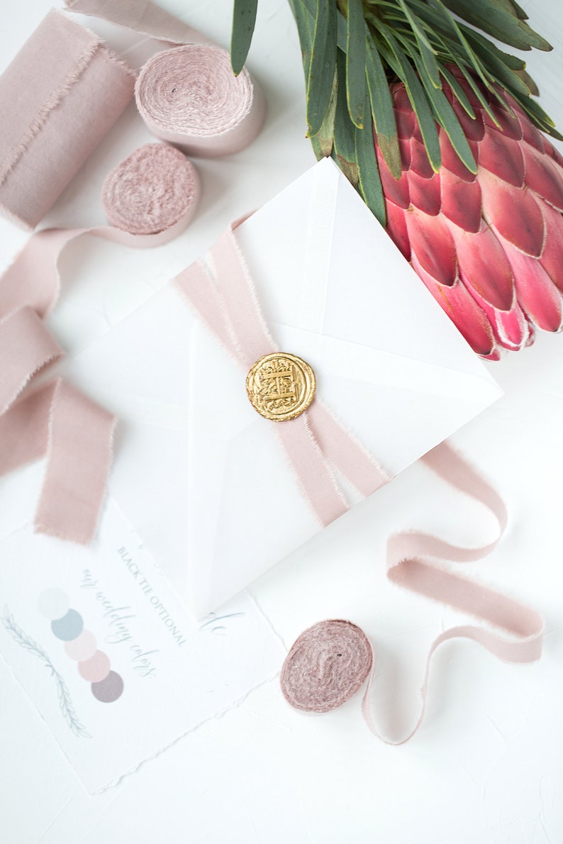 May include: A white envelope with a pink ribbon and a gold wax seal. The envelope is on a white surface with other pink ribbons and a pink flower.