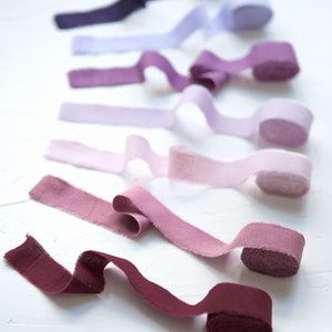 May include: A collection of eight fabric ribbon rolls in various shades of purple, pink, and red. The ribbons are arranged in a gradient from dark purple to burgundy.