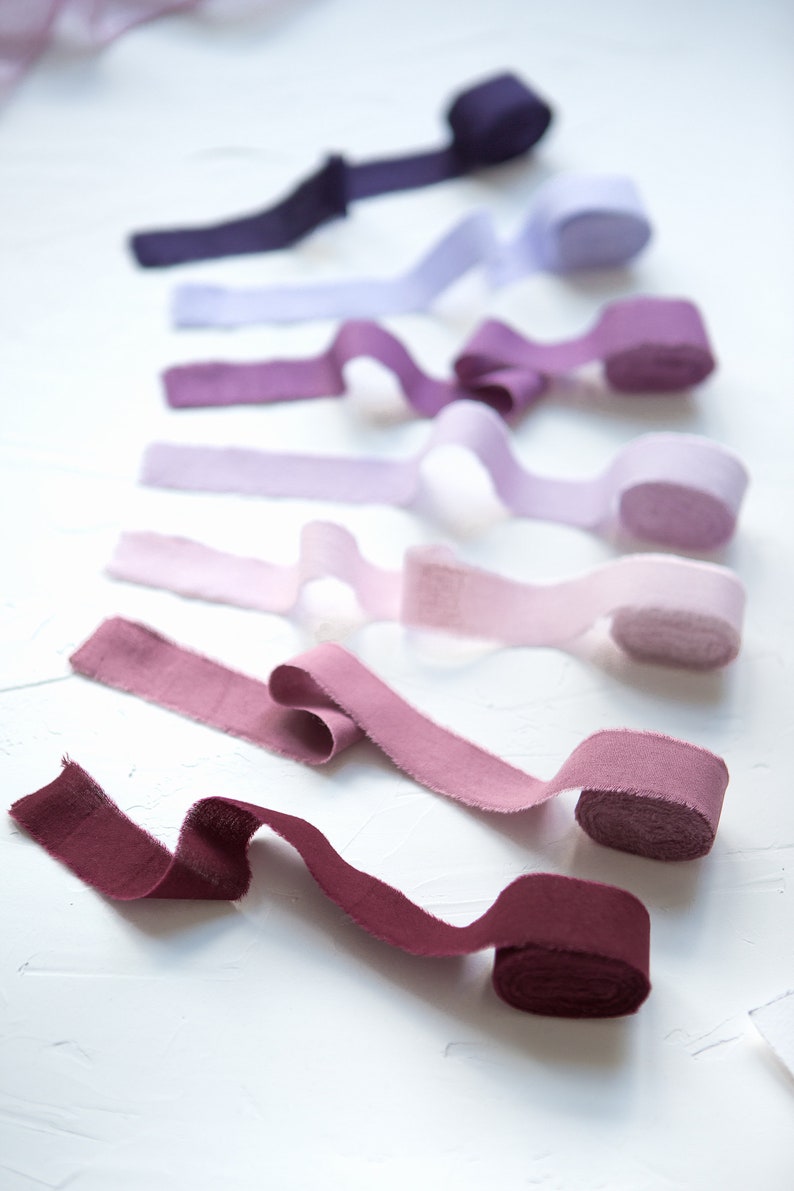May include: A collection of eight fabric ribbon rolls in various shades of purple, pink, and red. The ribbons are arranged in a gradient from dark purple to burgundy.