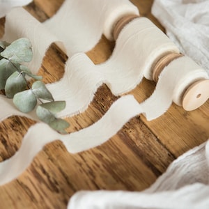 May include: A spool of white, frayed ribbon with a wooden dowel, on a wooden surface with a white fabric background.