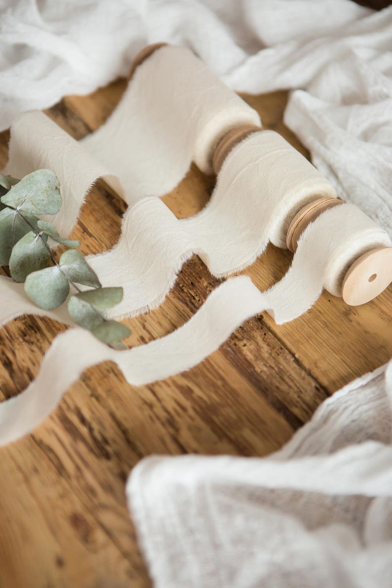 May include: A spool of white, frayed ribbon with a wooden dowel, on a wooden surface with a white fabric background.