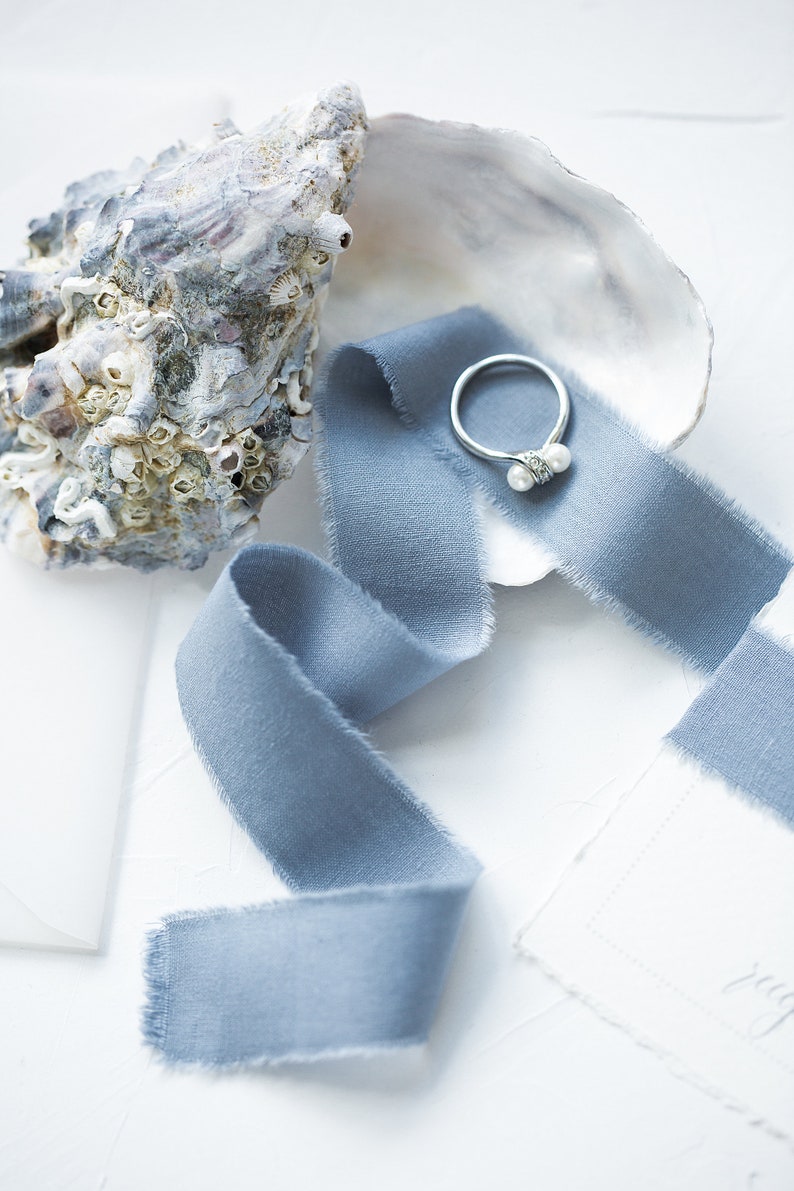 May include: A silver ring with two white pearls set in a seashell with a blue ribbon.