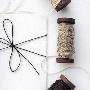 Może przedstawiać: Martwa natura z szpulkami sznurka i białą kartką związaną czarnym sznurkiem. Szpulki mają ciemne drewniane podstawy i są wypełnione naturalnym i brązowym sznurkiem. Karta jest biała i ma fakturę.