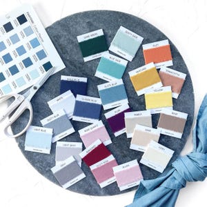 Op de afbeelding: Een verzameling stoffen stalen in diverse kleuren, waaronder donkergroen, dusty sage en roest, gerangschikt op een grijze stenen ondergrond. Een Pantone kleurenwaaier en een schaar zijn ook aanwezig. De kleuren zijn benoemd.