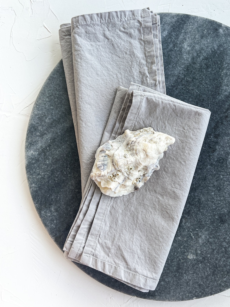 May include: Two gray linen napkins folded on a round gray marble surface. A white seashell is placed on top of the napkins.