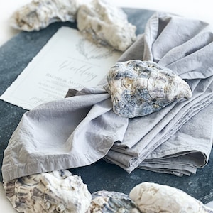 Pu&ograve; includere: Un tovagliolo di lino grigio con un anello per tovagliolo in conchiglia di ostrica naturale. Il tovagliolo &egrave; piegato e posizionato su una superficie di ardesia grigio scuro con un invito di nozze e altre conchiglie di ostriche.