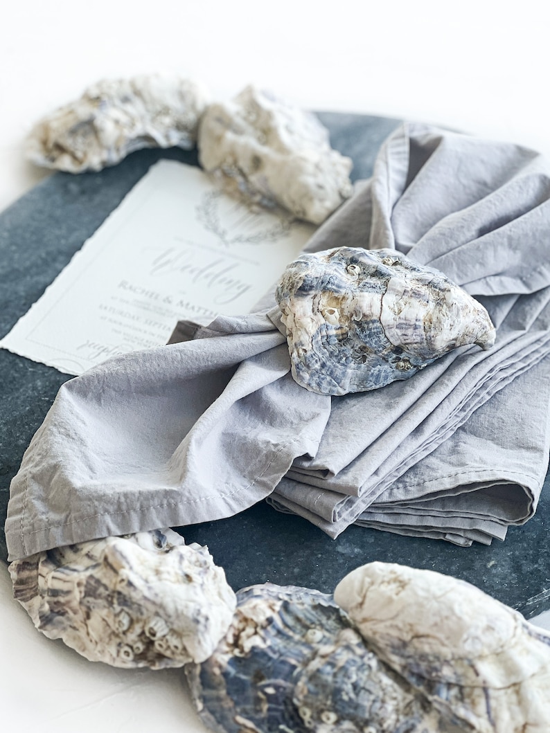 Pu&ograve; includere: Un tovagliolo di lino grigio con un anello per tovagliolo in conchiglia di ostrica naturale. Il tovagliolo &egrave; piegato e posizionato su una superficie di ardesia grigio scuro con un invito di nozze e altre conchiglie di ostriche.