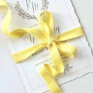May include: A wedding invitation tied with a yellow ribbon bow. The invitation card is white with black text, including the names "Rachel & Andrew" and the date "Saturday, S". The ribbon has a frayed edge.