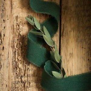 May include: A green ribbon with frayed edges is wrapped around a branch of green leaves on a rustic wooden background.