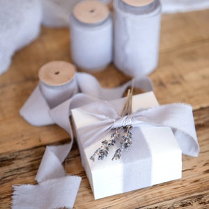 May include: A white gift box with a gray and white striped design is tied with a light gray ribbon and decorated with dried lavender sprigs.