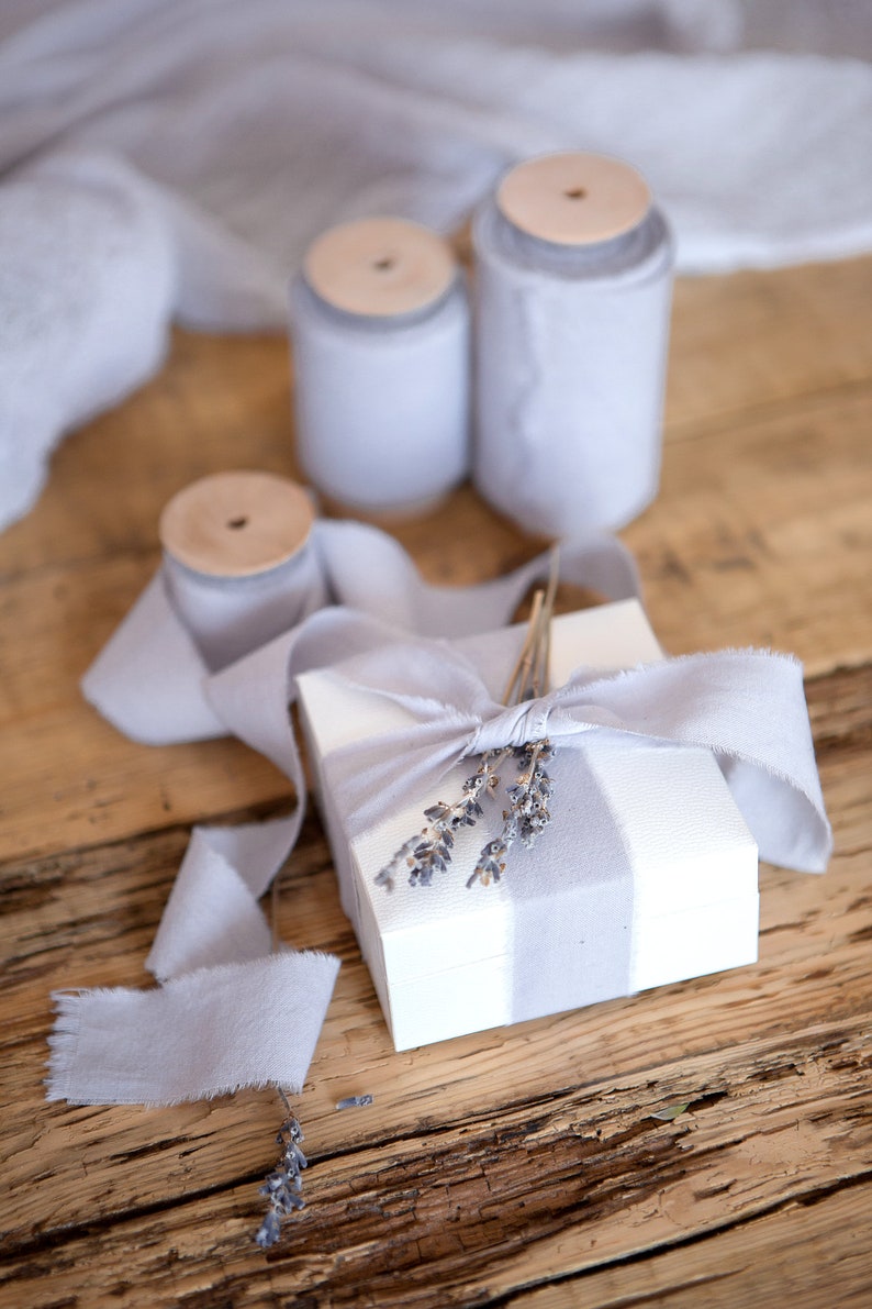 May include: A white gift box with a gray and white striped design is tied with a light gray ribbon and decorated with dried lavender sprigs.