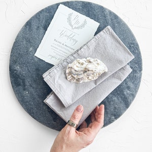 Pu&ograve; includere: Un invito di nozze con un carattere calligrafico su sfondo azzurro chiaro con una ghirlanda di foglie. L'invito &egrave; su un tovagliolo grigio con una conchiglia sopra. Il tovagliolo &egrave; su un piatto rotondo grigio.