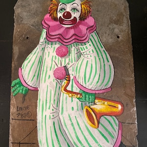 Op de afbeelding: Handgeschilderd clownkunstwerk op een leistenen tegel. De clown draagt een groen en wit gestreept pak, een roze kraag met ruches en een rode neus. De clown houdt een gouden saxofoon vast. Het kunstwerk is gesigneerd "LAICHE 783©".