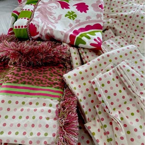 May include: A collection of bedding items featuring various patterns and colors. Includes pillowcases with floral and geometric designs in pink, green, and white, along with sheets adorned with pink and green polka dots. Some pieces have decorative fringe.