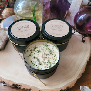 Puede incluir: Dos latas de velas de metal negro con tapas doradas y una vela blanca con motas verdes y marrones en su interior. Las latas de velas tienen una etiqueta que dice "Ankh of Anubis, Deluxe Collection, Apothecary".