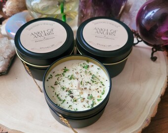 May include: Two black metal candle tins with gold lids and a white candle with green and brown flecks inside. The candle tins have a label that reads "Ankh of Anubis, Deluxe Collection, Apothecary".