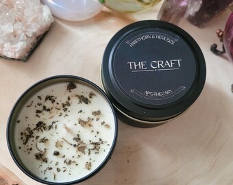 May include: Two black metal tin candles, one open and one closed, on a wooden surface. The open candle has a white wax with dried herbs. The closed candle has a black label with the text "THE CRAFT APOTHECARY".